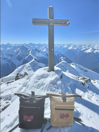 Zwei Rucksäcke stehen vor einem Kreuz auf schneebedecktem Berggipfel mit Alpenblick.