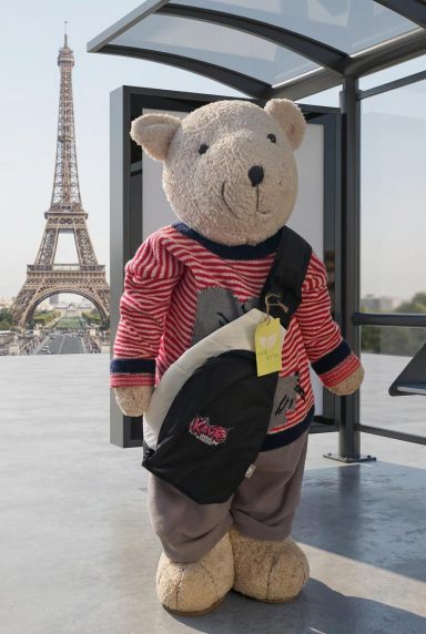 Ein großer Teddybär mit Tasche steht vor dem Eiffelturm.