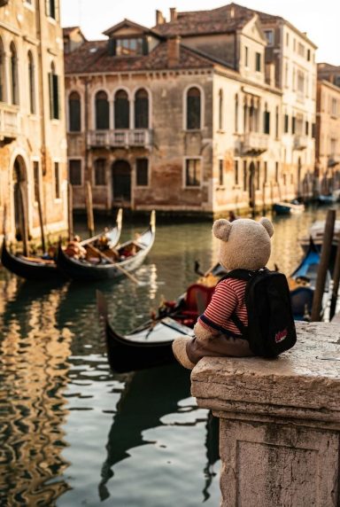 Ein Teddybär mit Rucksack sitzt an einem Kanal in Venedig, Gondeln im Hintergrund.